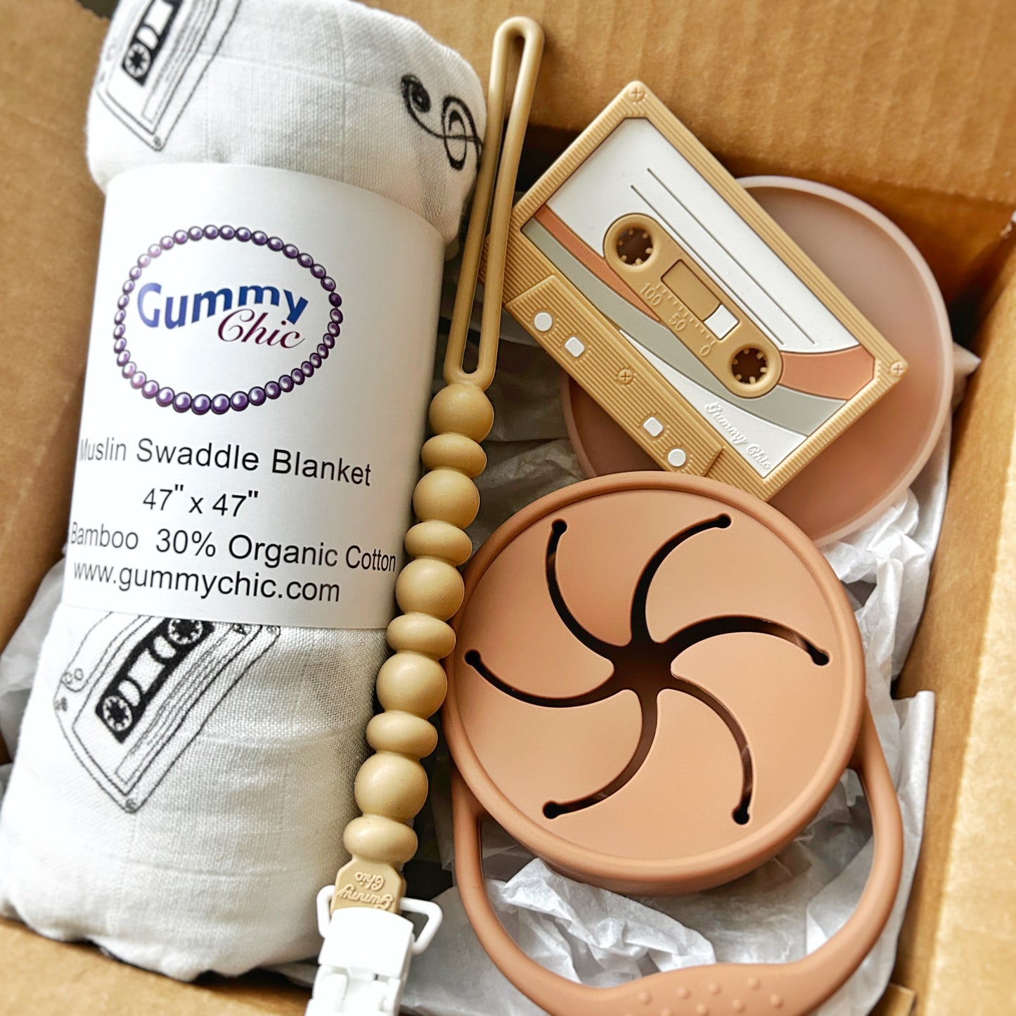 A gift box open to reveal a beige baby blanket with branding, a cassette tape teether, a silicone teether in progress, and a silicone collapsible snack cup.