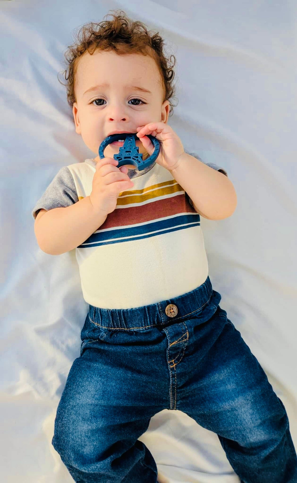Baby holding a Paris silicone teething toy, wearing a vintage-inspired outfit, sitting on a soft surface.