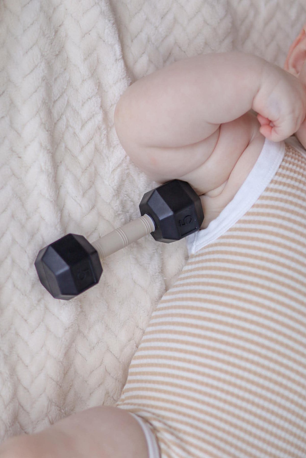 Baby lying on a blanket with a silicone dumbbell rattle teether toy beside them, showcasing a playful fitness theme.