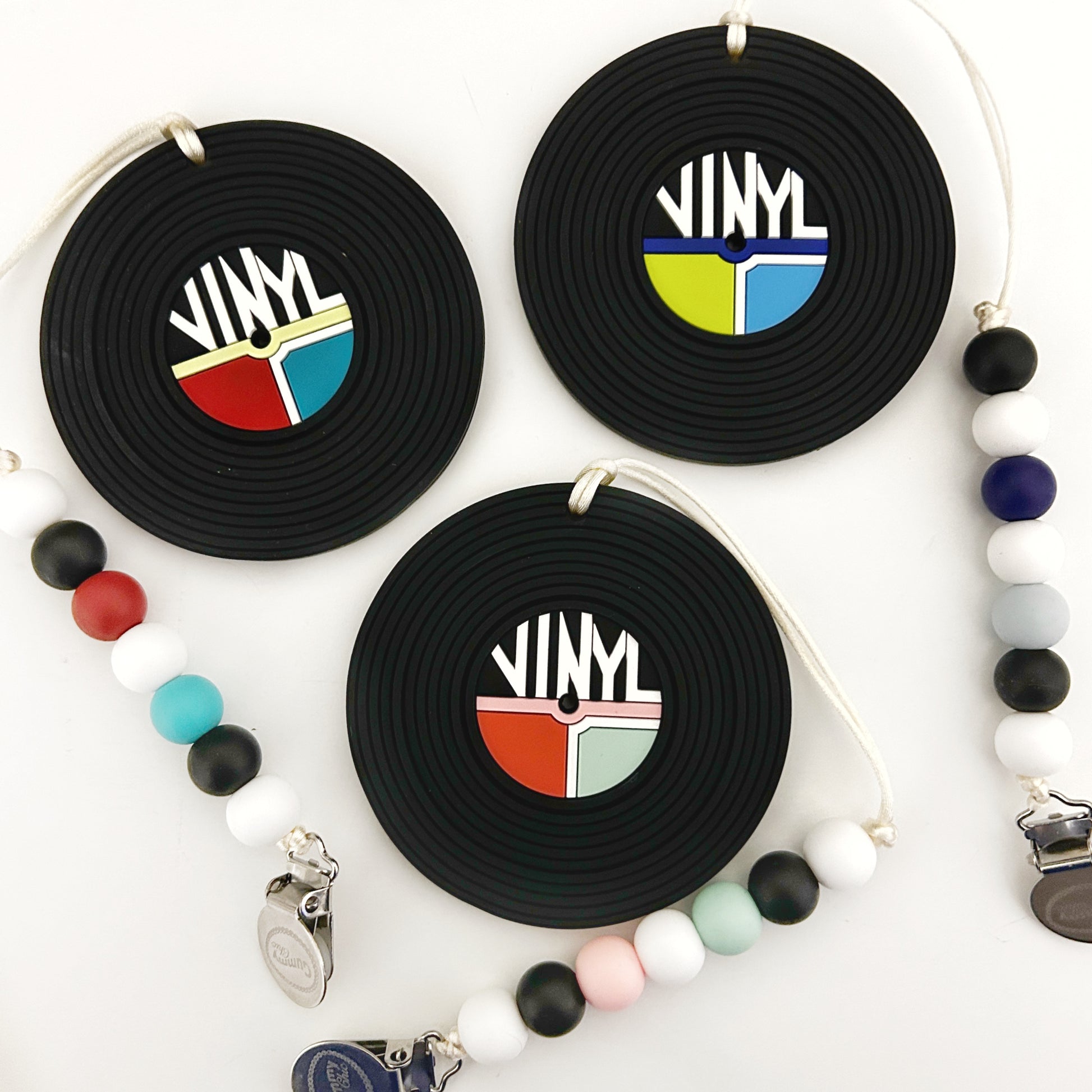 A selection of silicone teether toys designed to look like vinyl records, with various colorful designs in the center, attached to a clip. Other silicone beads and clips are also visible in the background.