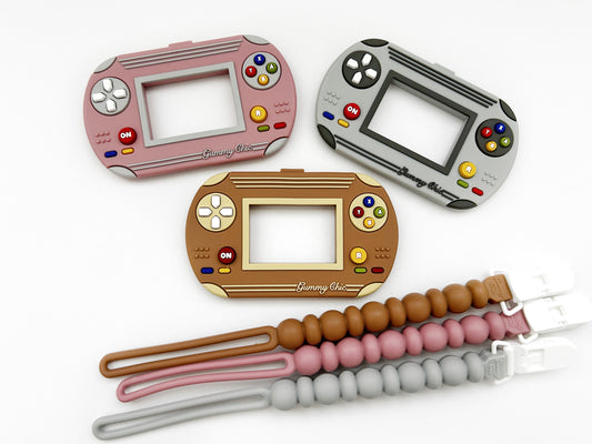 A silicone teether designed to resemble a video game console, in pink, gray, and brown colors, with beads attached to a clip.