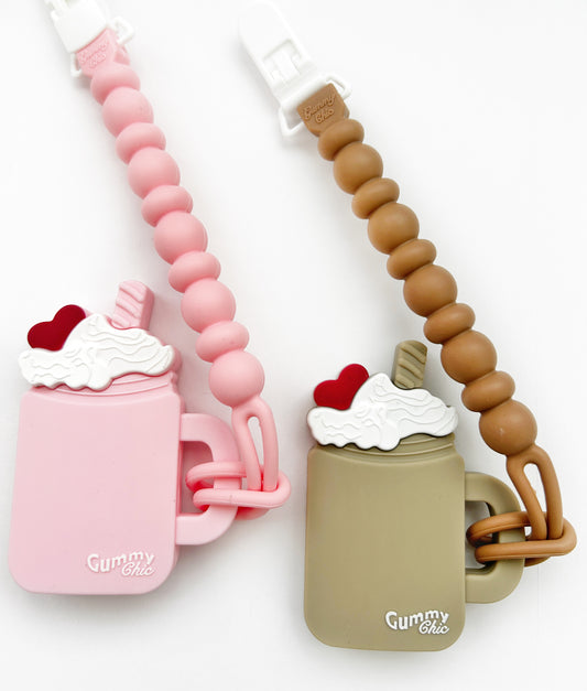 Two silicone teething toys designed to look like milkshakes with straws, one in pink with a chocolate design and the other in brown with a strawberry design. Each has a clip attached for attaching to strollers or clothing.