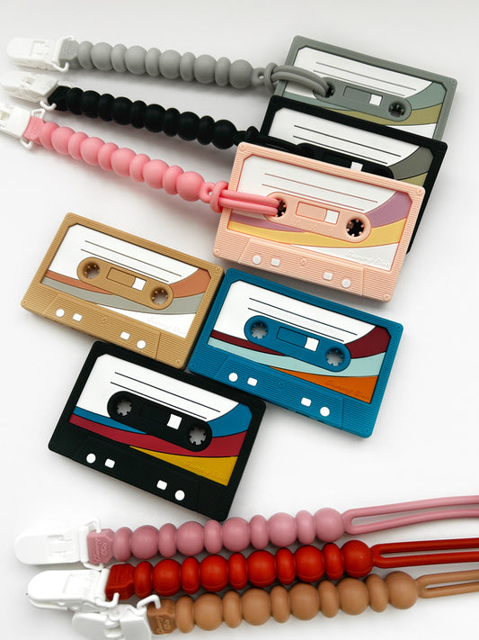 A collection of cassette tape-shaped teethers in various colors, designed to look like retro mix tapes. They are laid out against a white background.