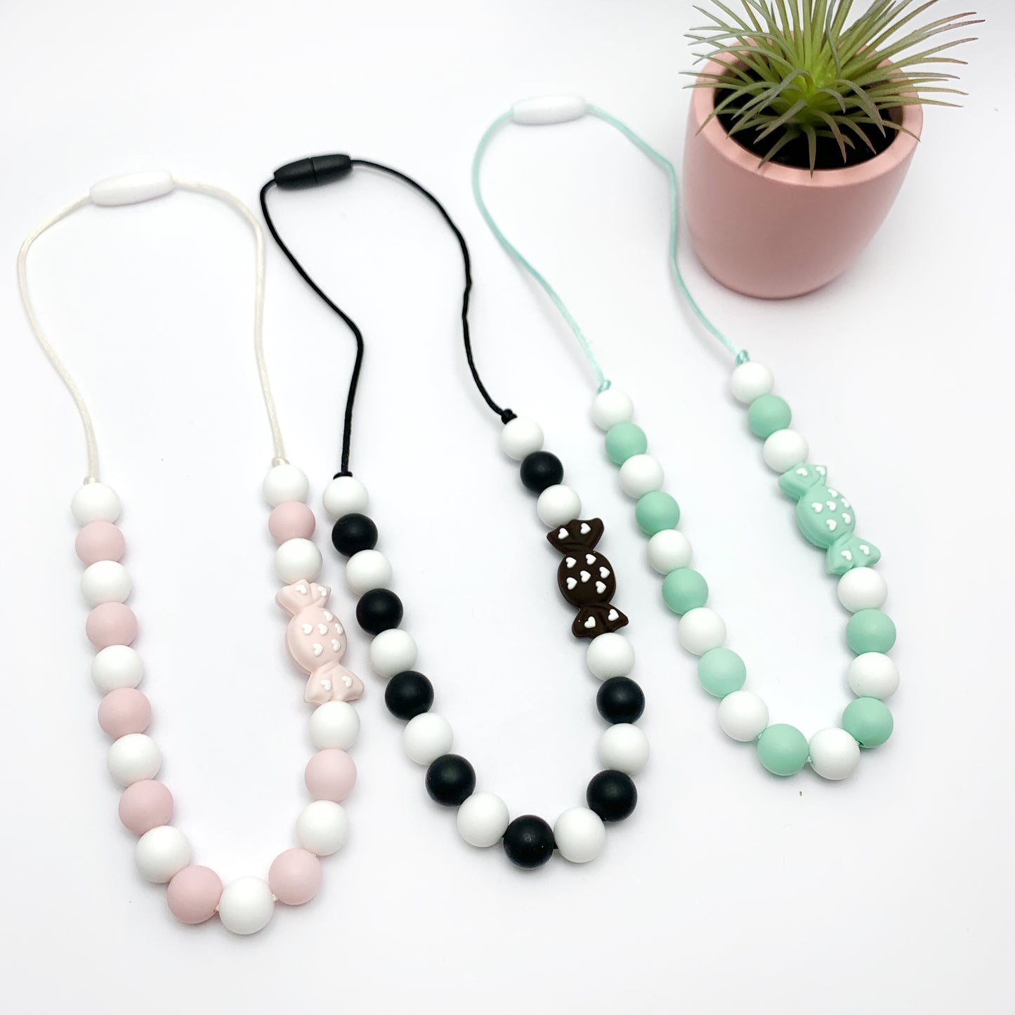 A collection of chewable sensory necklaces with silicone beads in various colors including black, white, pink, and green, displayed alongside a potted plant. The accent bead is a candy wrapper lookalike