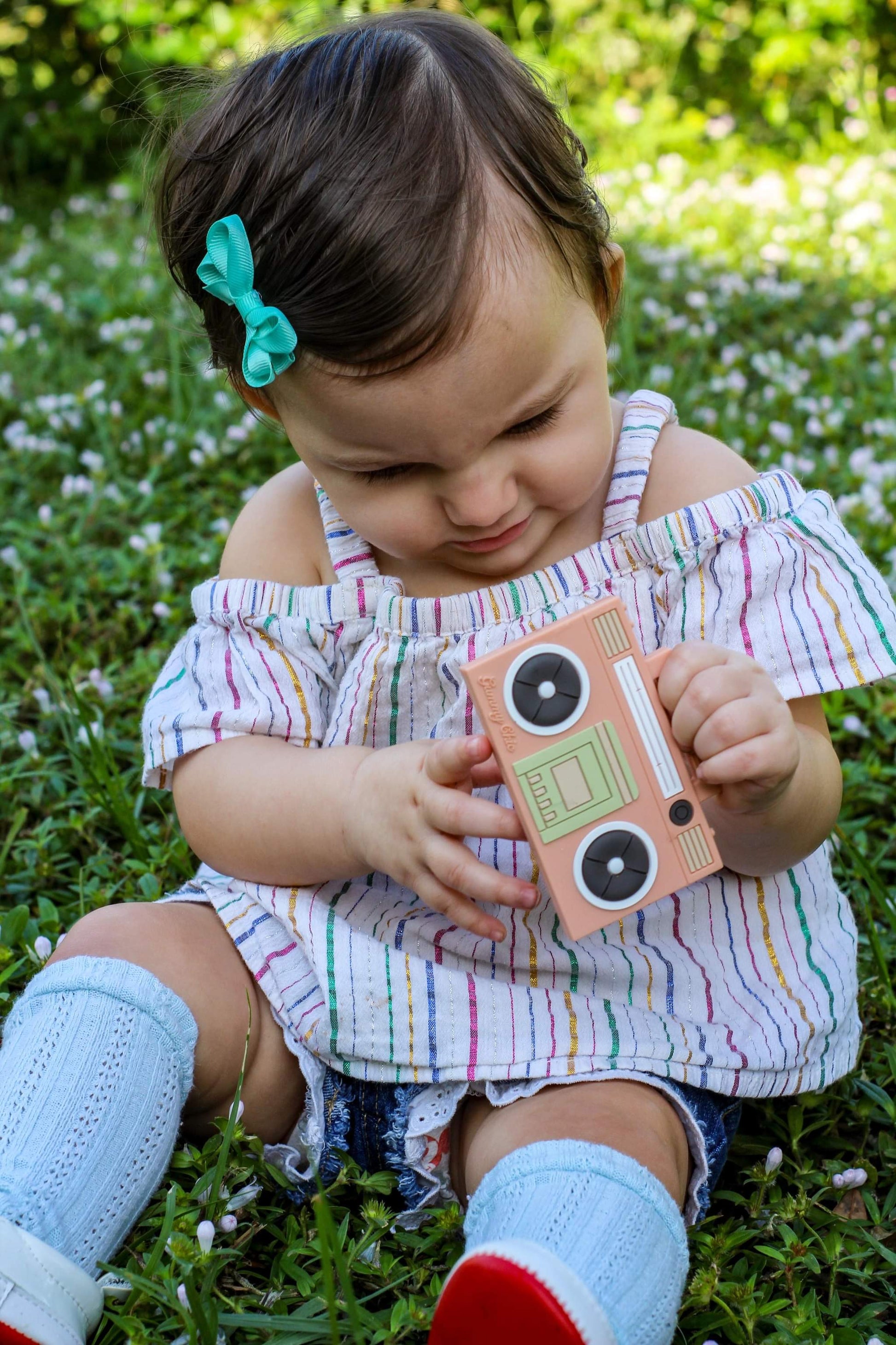 baby girl holding a tan silicone boom box teether sitting on the grass