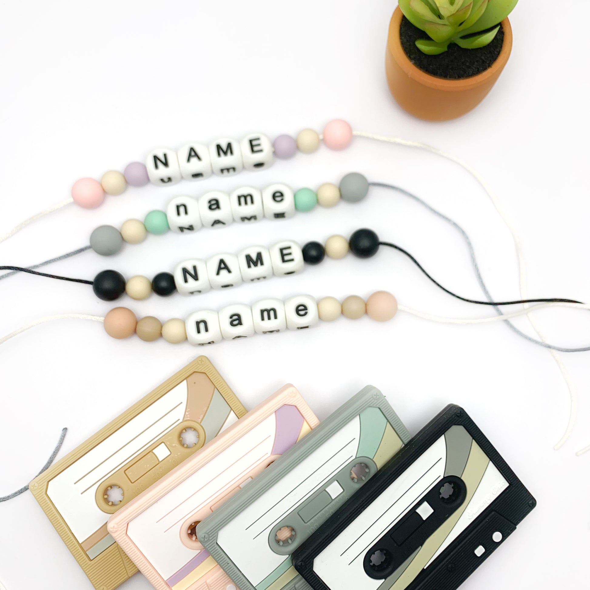 A collection of cassette tape-shaped baby teethers in various colors, designed to resemble retro cassette tapes. They are displayed alongside beads with names on them and a small potted plant.