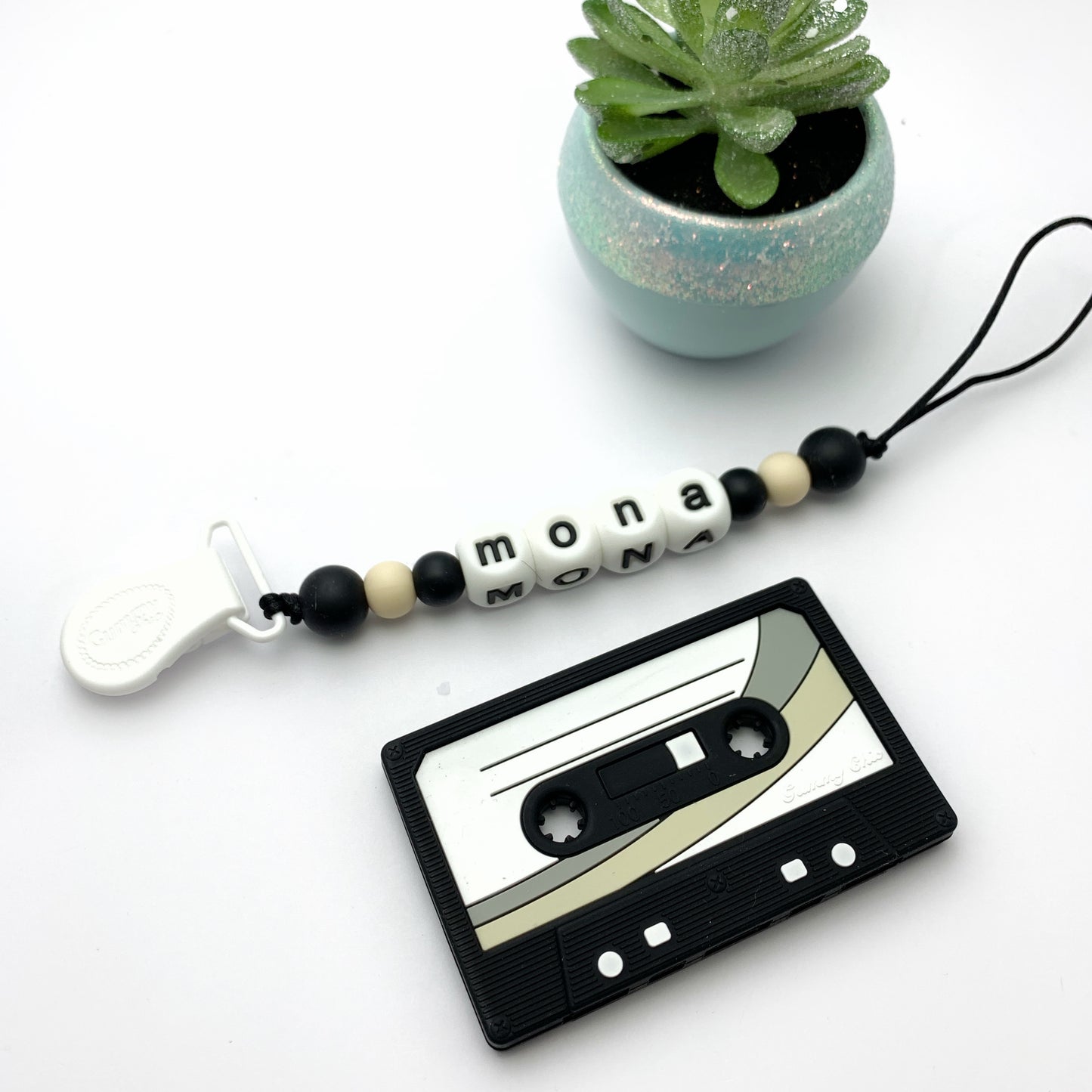A collection of cassette tape-shaped baby teethers in various colors, designed to resemble retro cassette tapes. They are displayed alongside beads with names on them and a small potted plant.