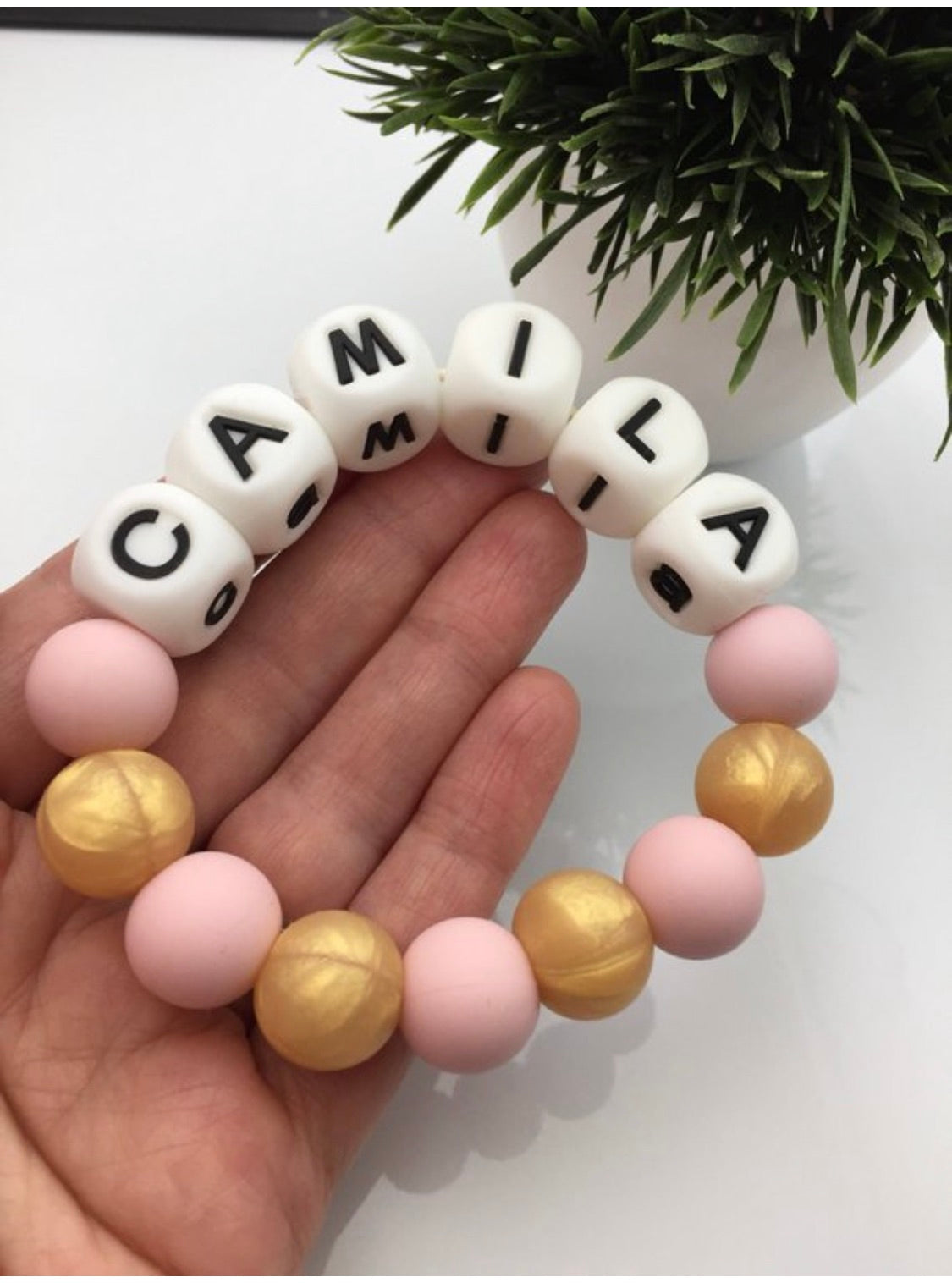 A hand holding a customizable teething ring made of silicone beads in various colors, with the name 'CAMILA' written on white beads.