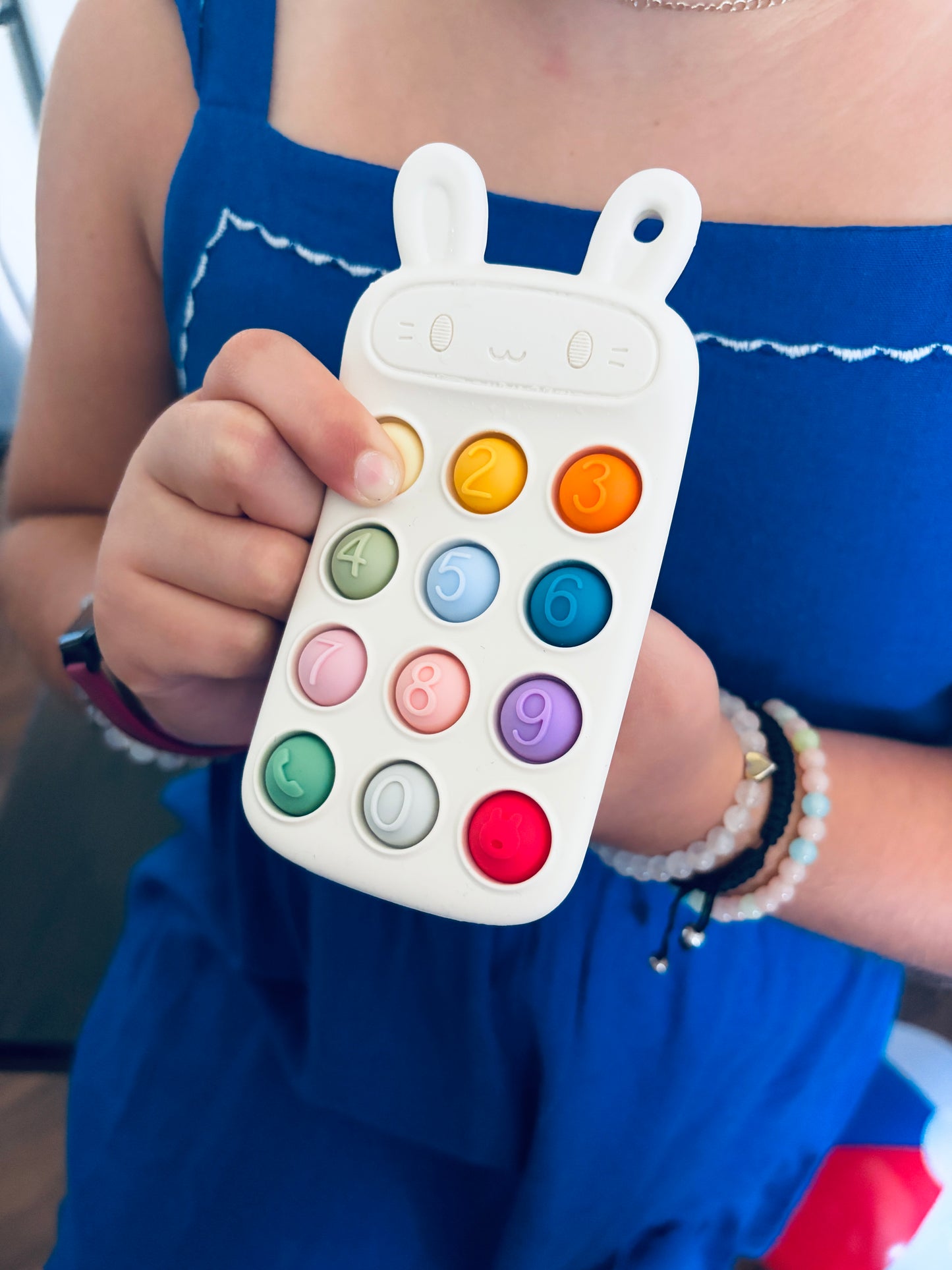 A close-up of a person holding a white silicone baby teething toy shaped like a rabbit head with colorful numbered buttons, wearing a blue dress.