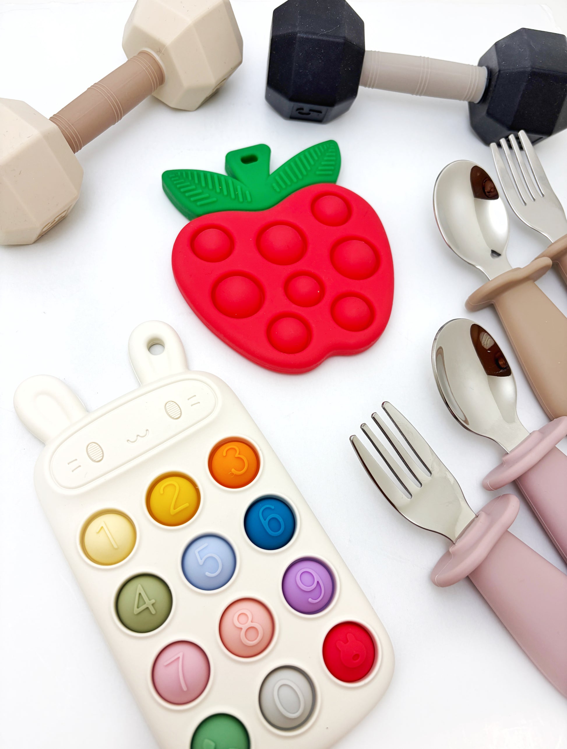 A styled flat-lay of baby feeding and sensory toys on a white background, including a red strawberry-shaped silicone teether with green leaves, a bunny-shaped number/activity learning toy, a black-and-beige dumbbell, and pastel-colored utensils with forks, spoons, and handles.
