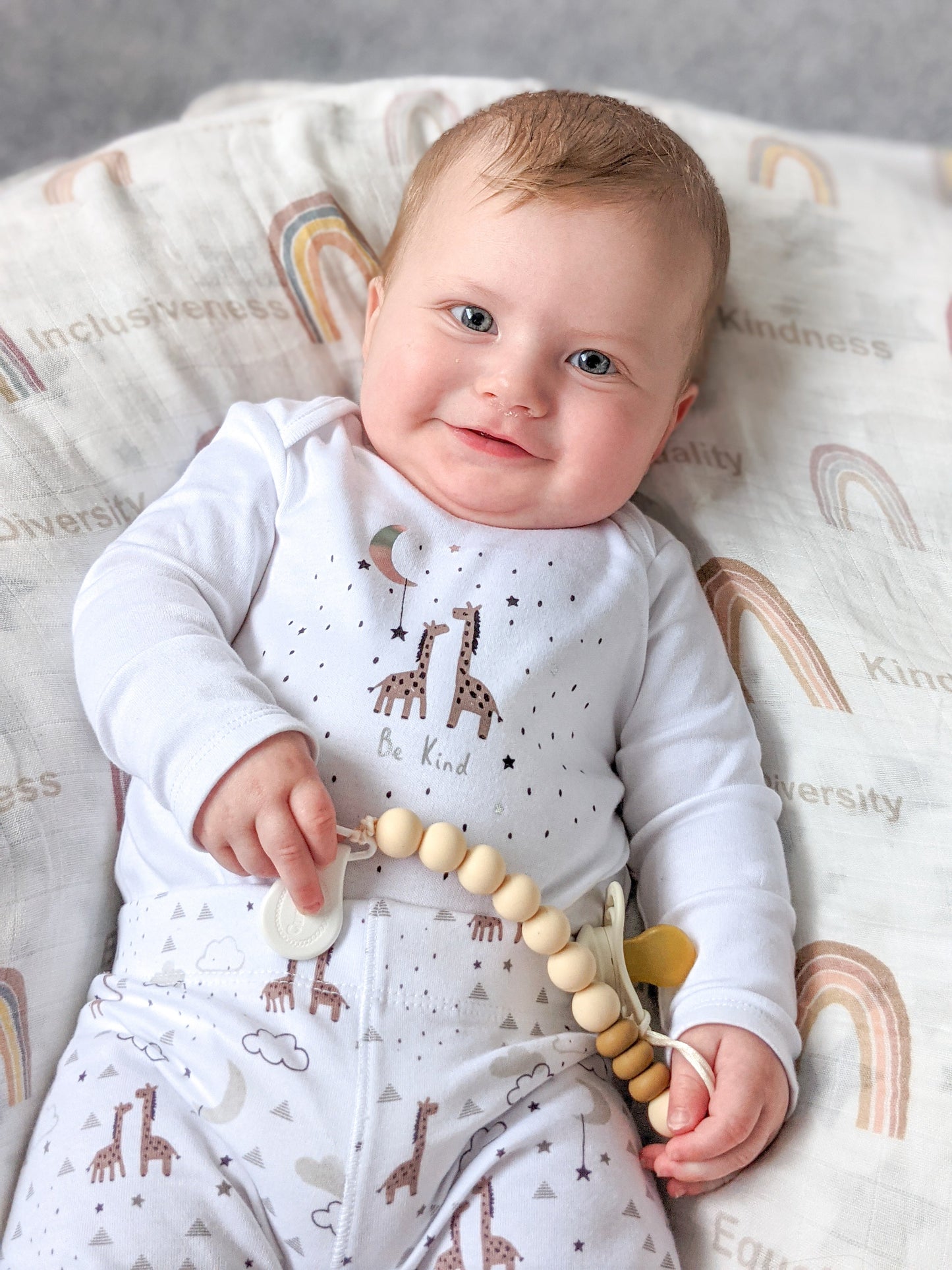 baby smiling holding a pacifier clip with a rainbow inclusive blanket on the back.