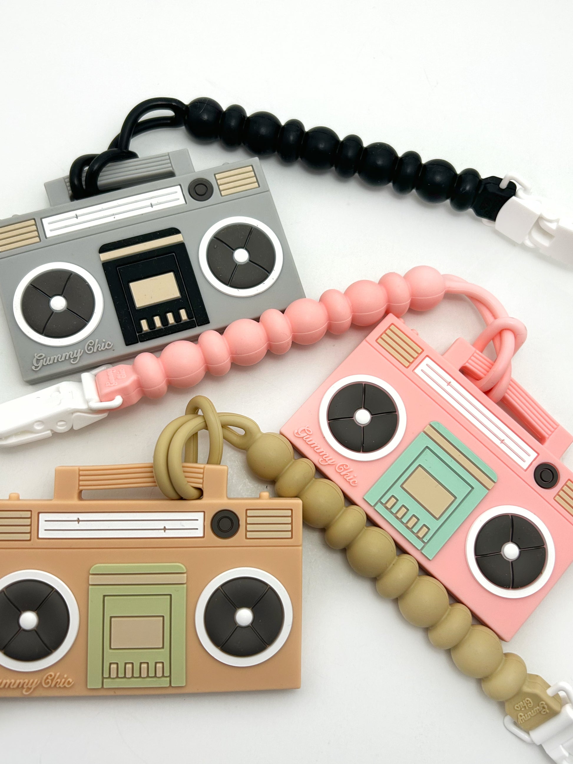 A silicone teether designed to look like a boom box, with a clip attached to it. The teether is pink with white and gray accents, and includes a clip-on feature for easy attachment to a stroller or other baby gear.