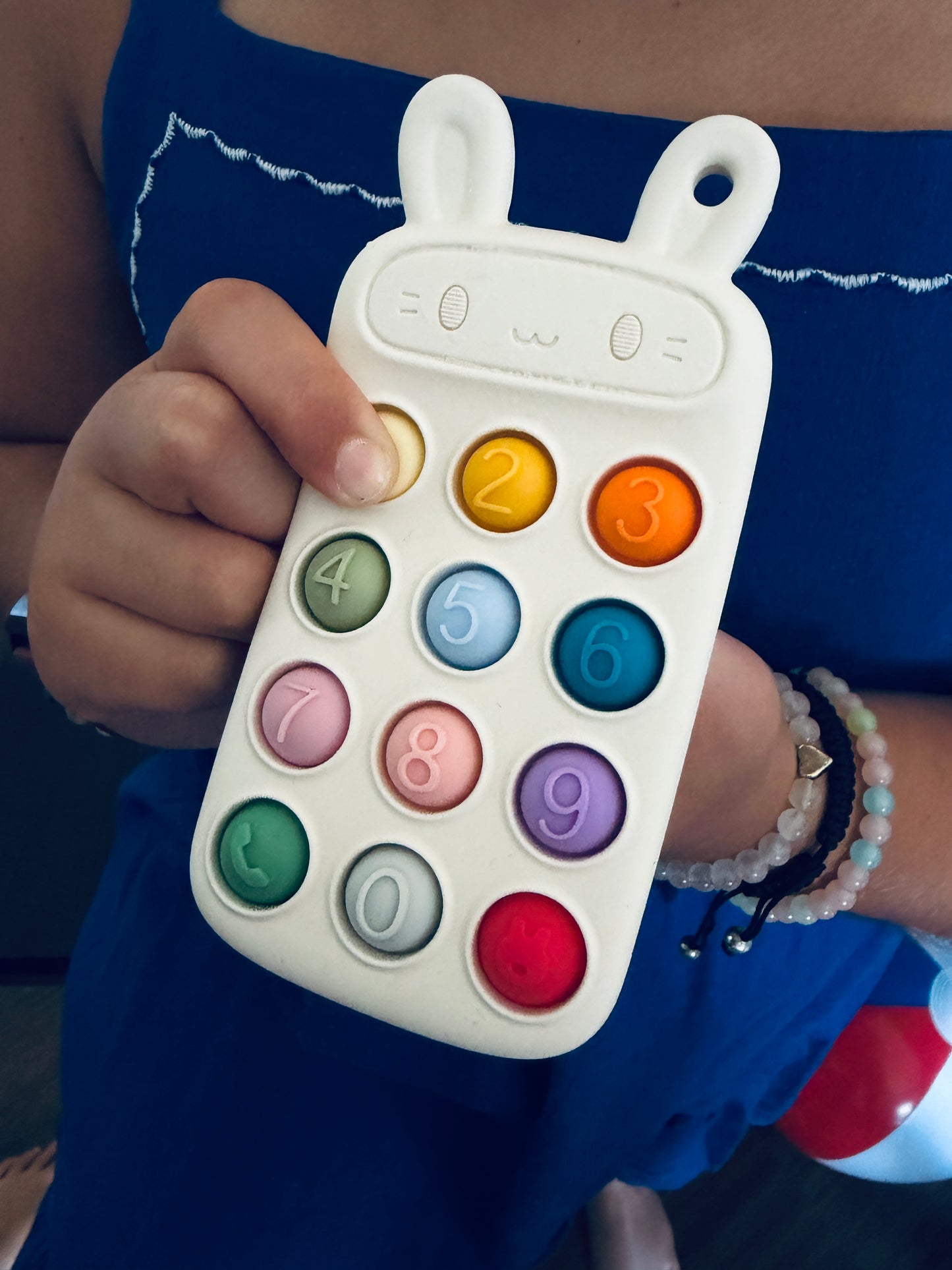 A baby toy calculator with bunny ears, held by a person wearing a blue dress. The calculator is white with colorful round buttons numbered 0 through 9.