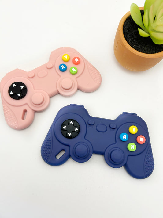 Two silicone teethers designed to resemble game controller pads, one in pink and one in blue, with colorful buttons, on a white surface next to a potted plant.