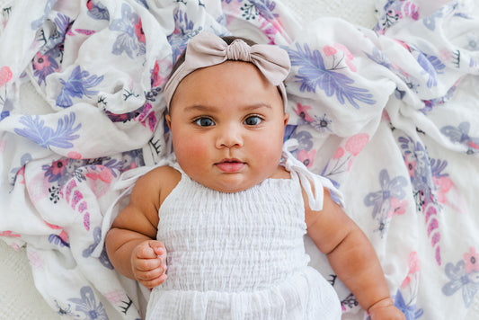 A baby lying on a floral muslin blanket with a focus on the baby's face. The baby is wearing a white outfit and has a pink bow on the head. The blanket is purple with unicorn figures and pink flowers
