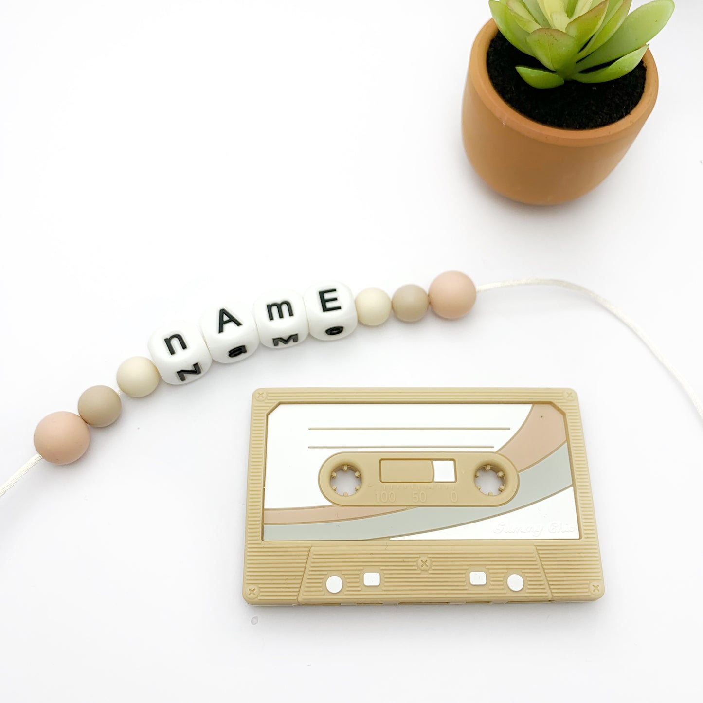 A collection of cassette tape-shaped baby teethers in various colors, designed to resemble retro cassette tapes. They are displayed alongside beads with names on them and a small potted plant.