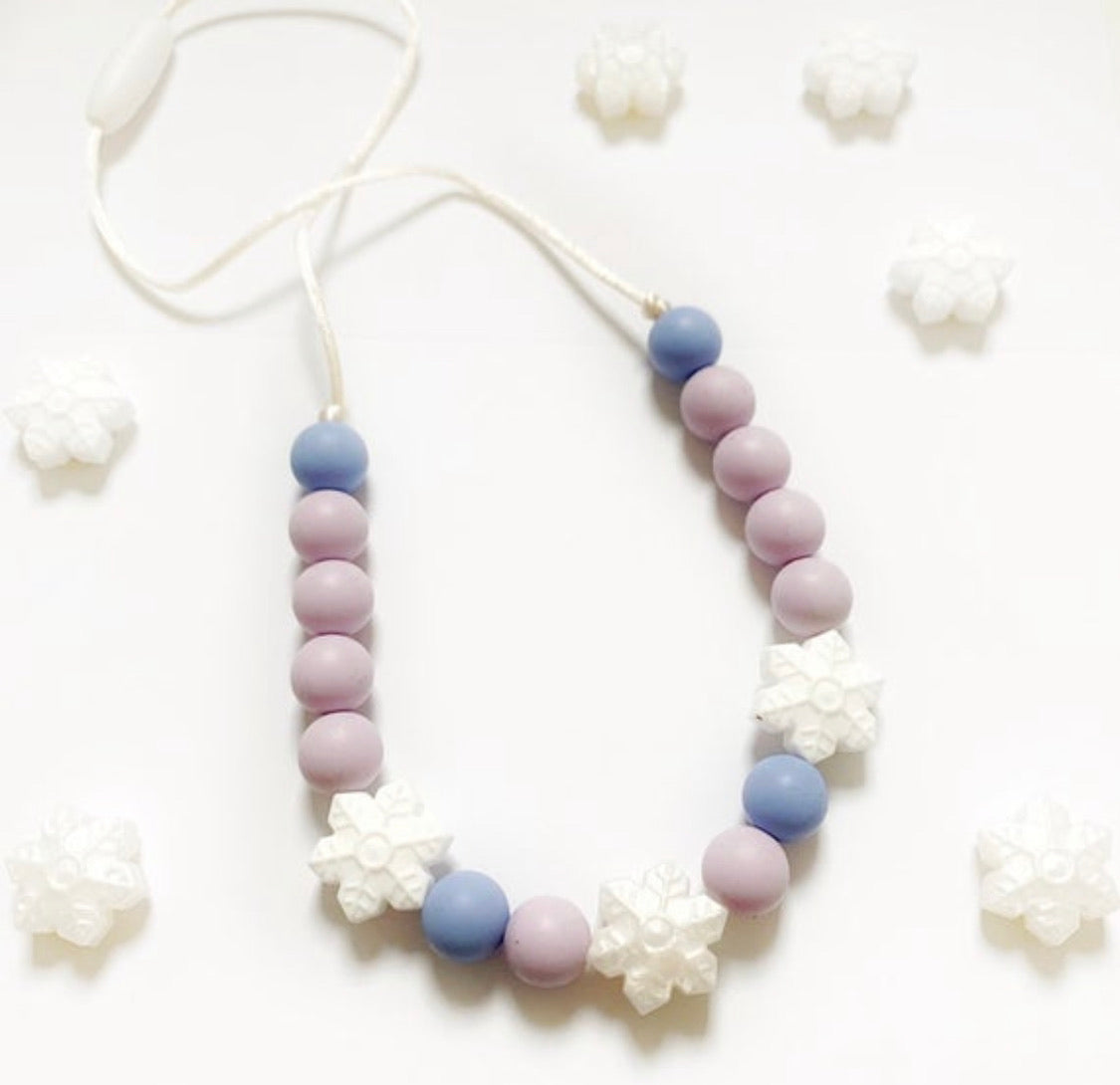 A necklace with purple, blue, and white beads, featuring white flower-shaped charms, displayed against a white background with additional white flowers.