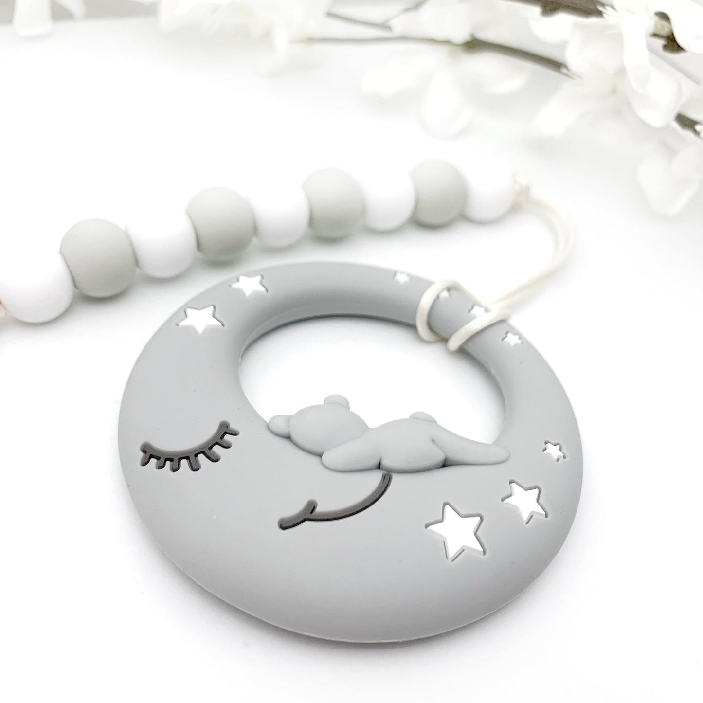 A gray teething toy in the shape of a moon with stars, attached to a clip with a white leash made of silicone material.
