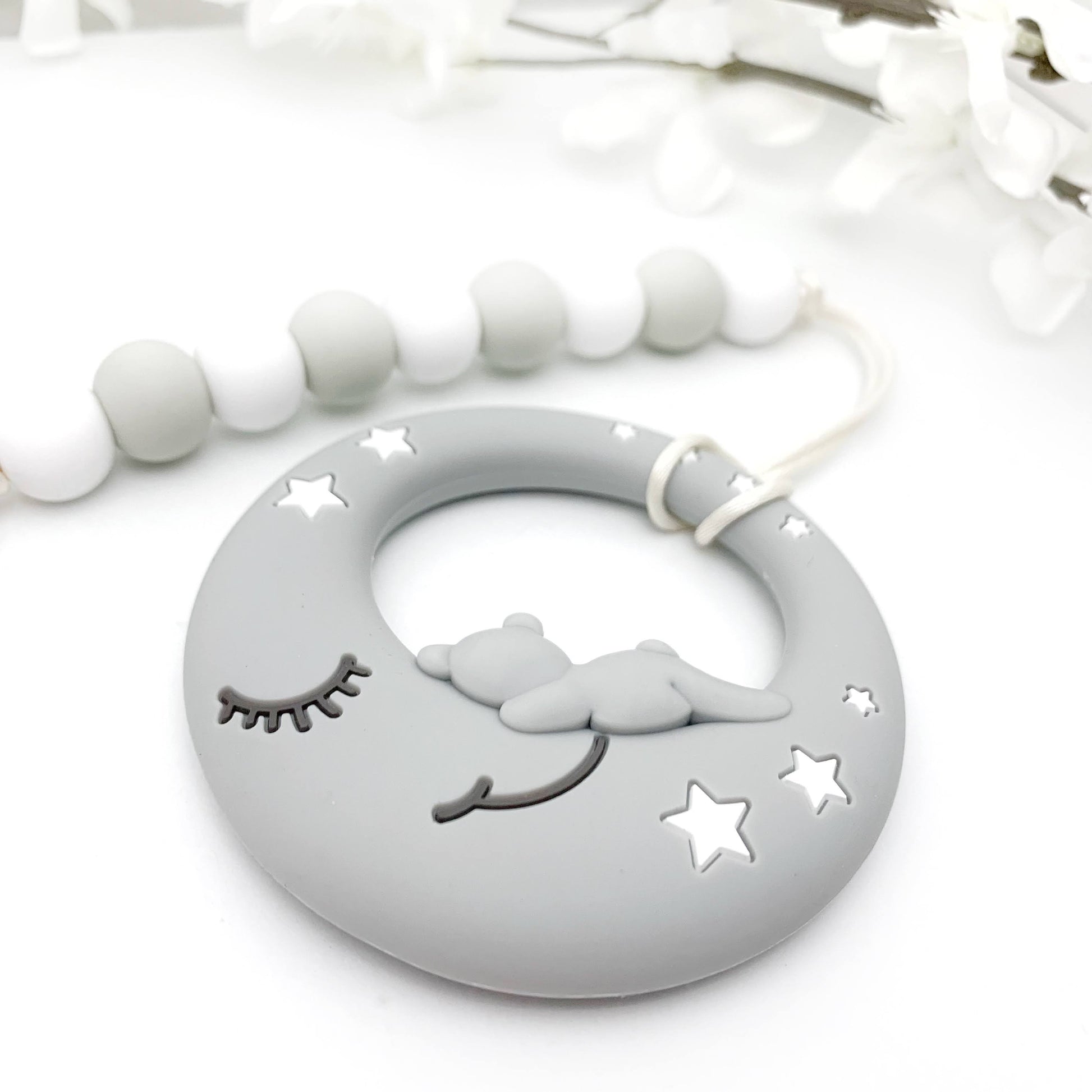 A gray teething toy in the shape of a moon with stars, attached to a clip with a white leash made of silicone material.