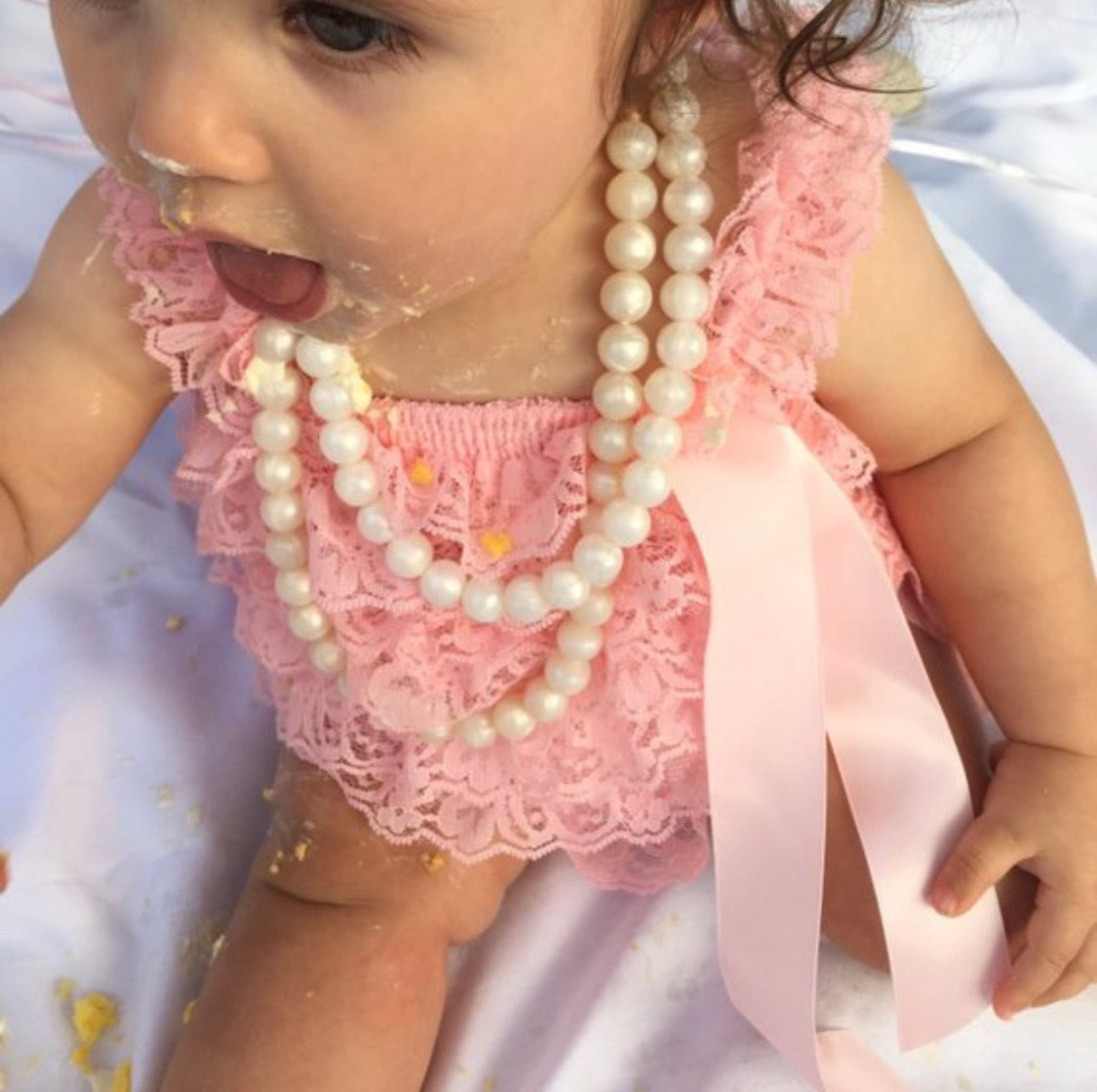 A baby wearing a pink lace outfit and two white silicone bead necklaces with a breakaway clasp.
