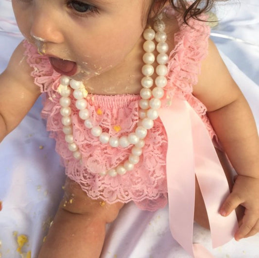 A baby wearing a pink lace outfit and two white silicone bead necklaces with a breakaway clasp.