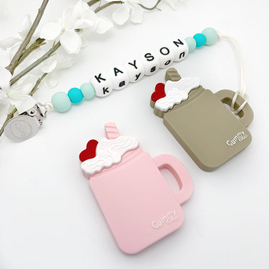 Silicone teething toy designed to look like a milkshake with a cherry on top, presented in a pink color. The toy has a clip attached to it and is displayed alongside a name bead chain.