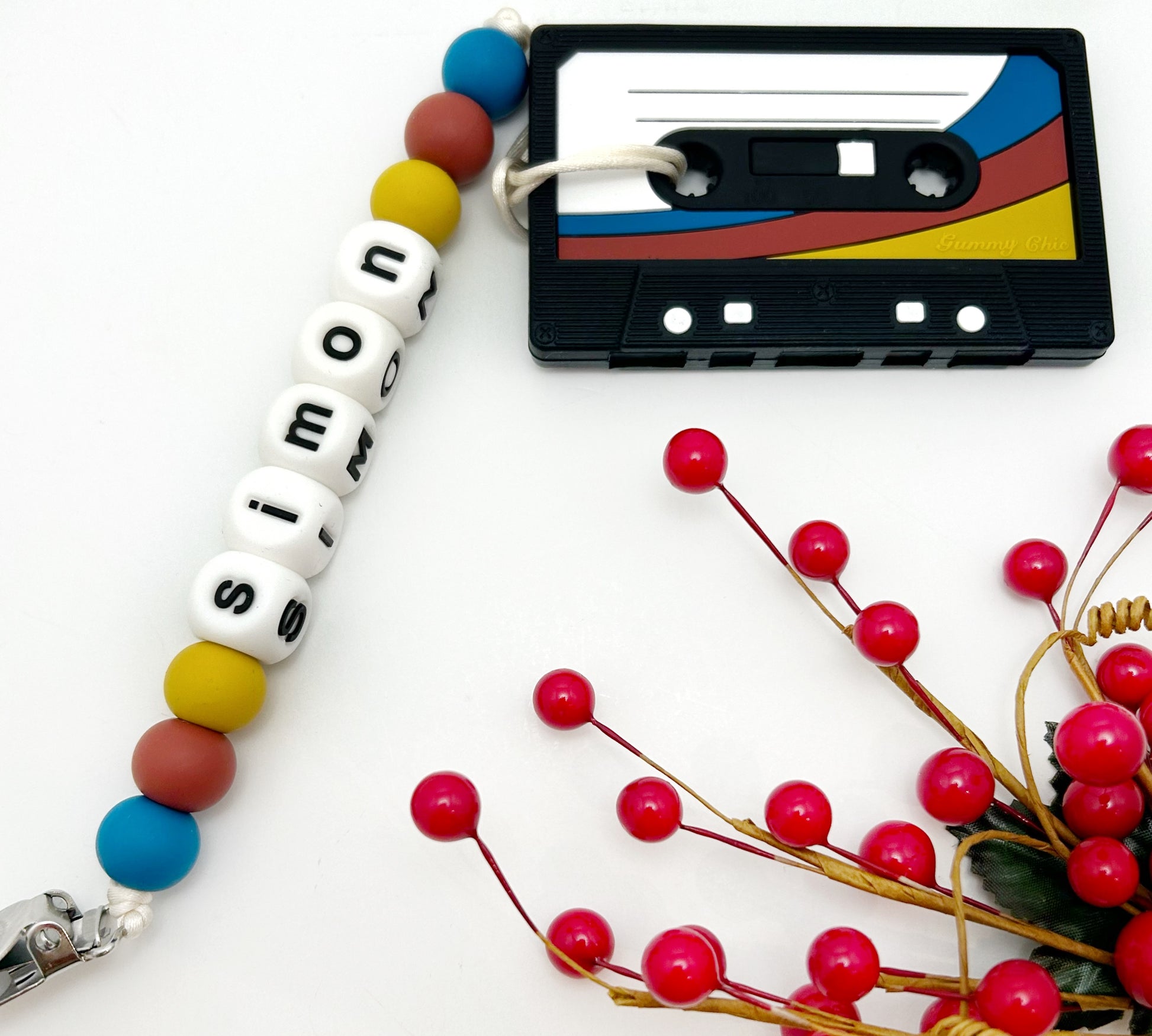 A teething toy designed to resemble a cassette tape, with multicolored beads on a strap. The cassette has a black background with red, blue, and yellow stripes.