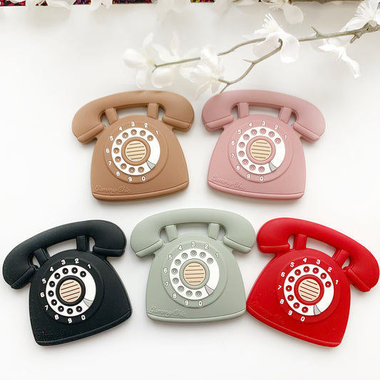A collection of Gummy Chic Cool Baby Teethers in various colors designed to resemble rotary dial phones. They are displayed in two rows with colors including beige, pink, gray, black, and red.