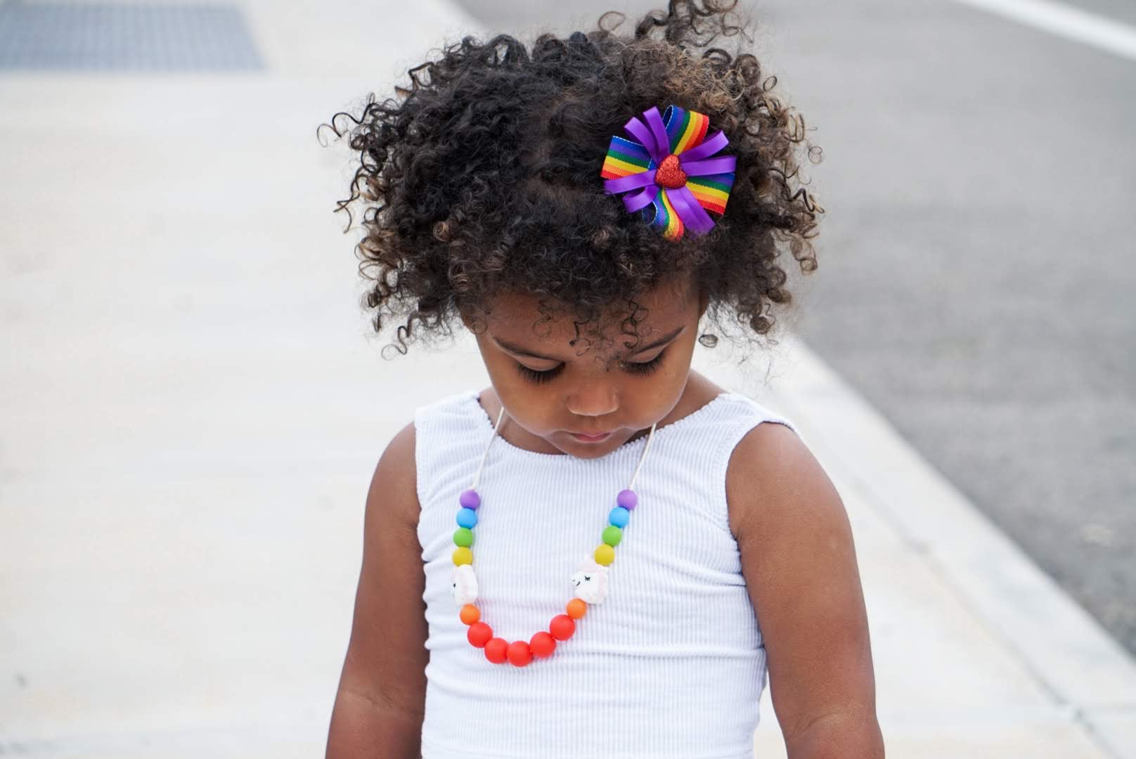 A child with dark curly hair is wearing a white sleeveless top and a silicone sensory necklace with vibrant rainbow colors.