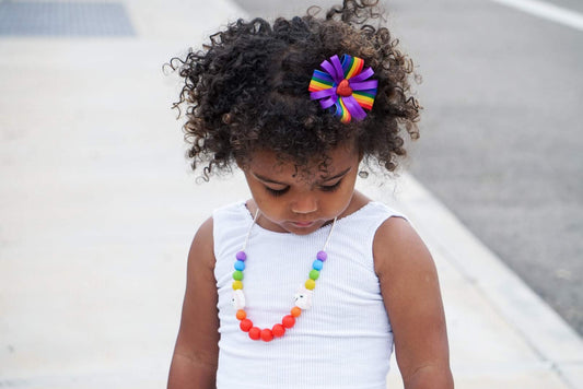 A child with dark curly hair is wearing a white sleeveless top and a silicone sensory necklace with vibrant rainbow colors.